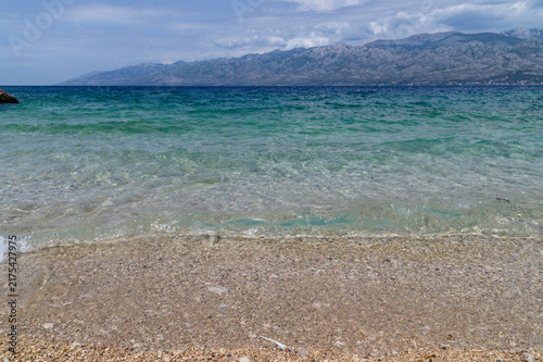 Crystal clear turquoise water over a pebble shore with barren karst mountains across the bay, Dalmatia, Croatia