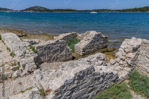 Crumbling Roman walls on the shoreline of the Colentum site with a village across the bay, Murter, Croatia