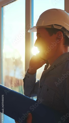 A man in a white hard hat speaks on the phone while holding a blueprint. The man is positioned near a large window, indicating a construction or renovation setting.