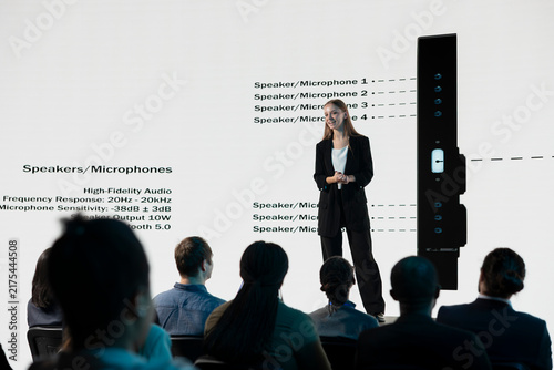 Young adult presents a mobile phone announcement on stage with promotional visuals, advertising a new release and investment deal during a business convention and product launch event for investors.
