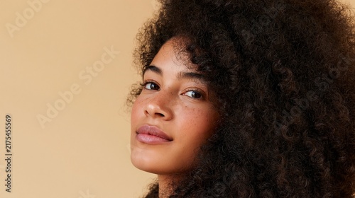 Looking confidently directly into the camera, a beautiful African American girl with natural curly afro hair and delicate freckles poses against a soft beige studio backdrop.