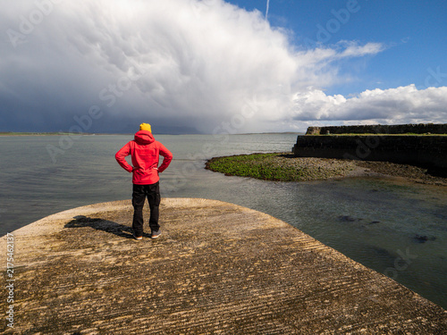 Принти на полотні Red-jacketed figure overlooks stormy sea from rocky outcrop, evoking solitude an