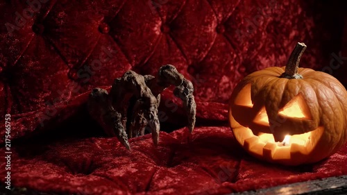 Spooky halloween jack o lantern pumpkin glowing next to a creepy monster hand crawling on a luxury red velvet tufted sofa couch background