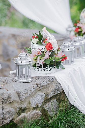 Konstfotografi Outdoor Celebration Decor: A charming white birdcage, adorned with vibrant pink and coral flowers, serves as a centerpiece on a stone wall, flanked by decorative lanterns and flowing white fabric