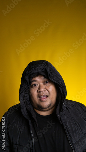 Fotografie An expressive young Asian male looks directly at the camera with a slightly open mouth while wearing a black winter jacket against a solid yellow background