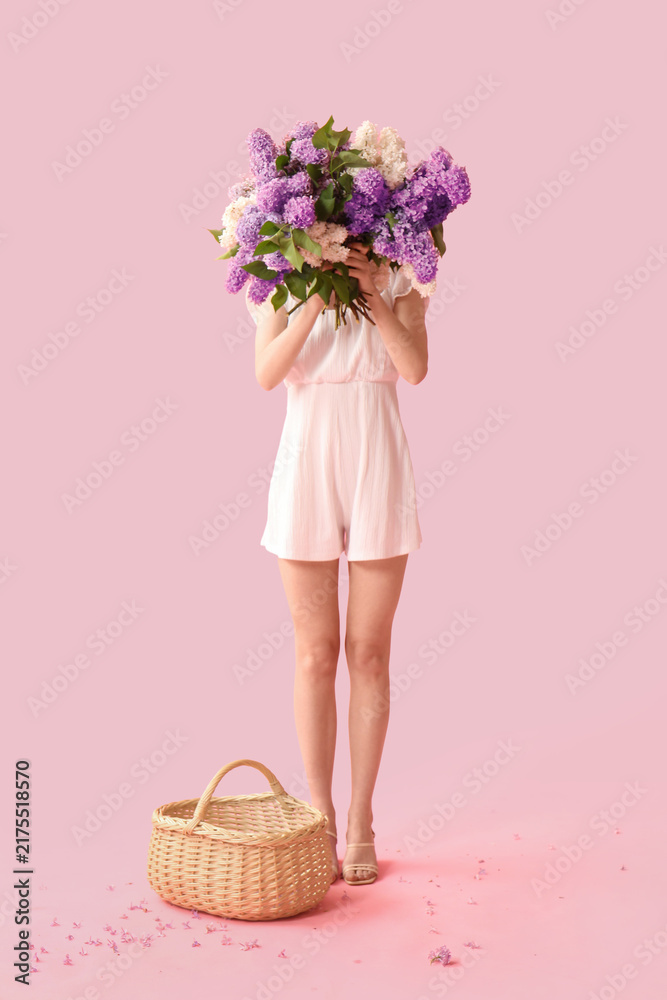 Obraz premium Beautiful young woman with wicker basket and bouquet of lilac flowers on pink background