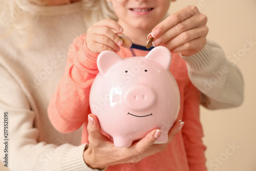 Cuadro en lienzo Little boy with his grandmother putting coins in piggy bank on beige background,