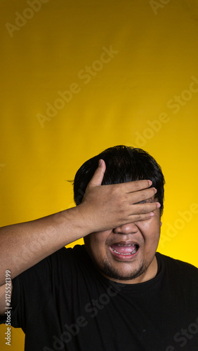Фотография A heavily stressed young Asian male firmly covers both eyes with one hand while keeping the mouth open in a distressed groan against a vibrant solid yellow background