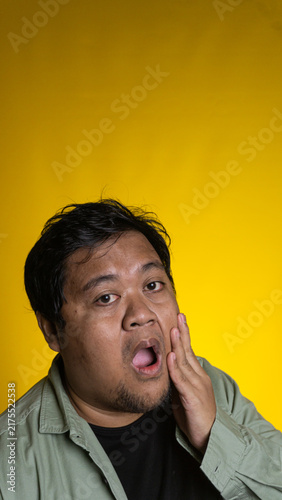 Konstfotografi A surprised young Asian male holds one hand against the cheek with a wide open mouth against a cheerful solid yellow background