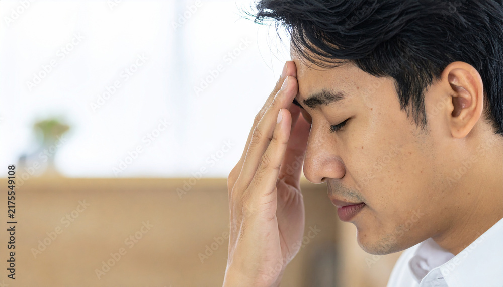 Fototapeta premium Headache and Stress: A young Asian man with his eyes closed touches his forehead, conveying a sense of discomfort and intense headache or mental fatigue.