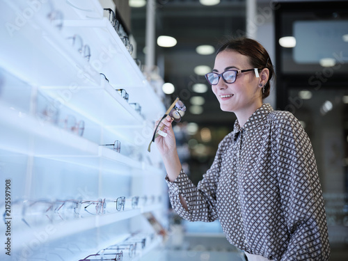 Foto Attractive White female customer, cheerful, chooses and tries on variety of eyeglasses for improved eyesight for individual lifestyles, makes decisions in options choices at display in optical shop