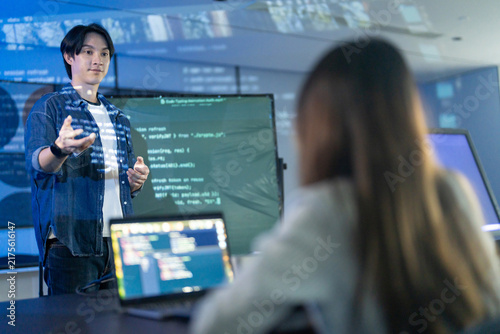 Asian male programmer giving coding presentations to IT team in the modern office. Tech startup developer explaining software on screens. Corporate innovation and digital technology business concepts
