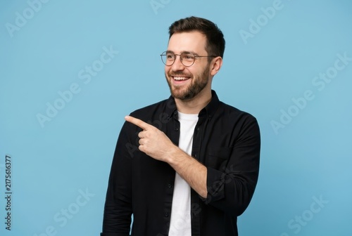 Happy Young Bearded Man in Glasses Pointing Finger Aside at Copy Space on Blue Background