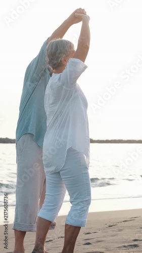 Cheerful senior couple holding hands and dancing barefoot on the wet sand of the beach at sunset, enjoying vacation and retirement with smiles, love and complicity in a moment of pure happiness