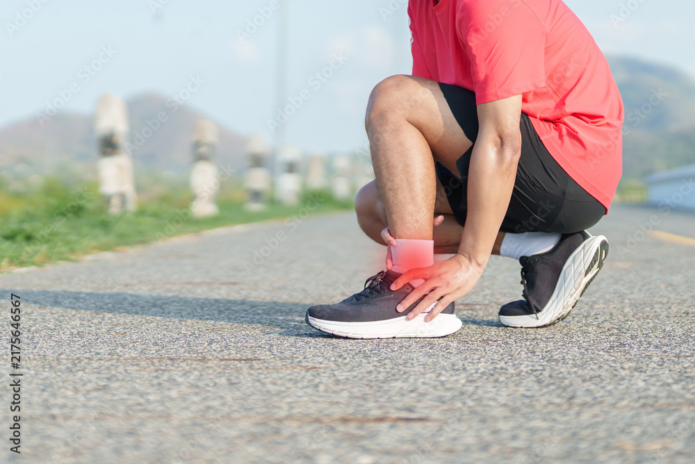 Obraz premium Young adult man suffering from acute ankle pain during running workout. male runner holding his leg due to sports injuries, ankle sprains or achilles tendonitis. Medical and healthcare concept