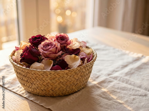 Dried roses in woven basket with petals on linen tablecloth by window, elegant home decor or aromatherapy concept.
