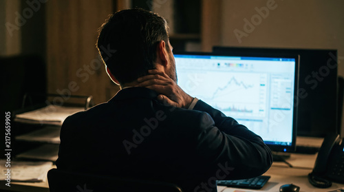 Man viewing financial chart on computer monitor while rubbing his neck in dark office, expressing fatigue during demanding workday