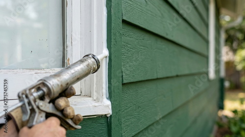 Sealing exterior window frame with caulk from gun on green house, home improvement and weatherization.
