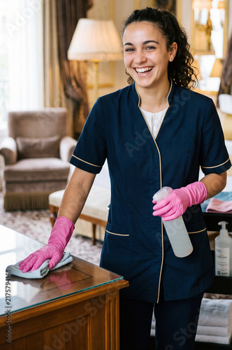 Smiling female housekeeper cleaning table with spray bottle and cloth in luxury hotel room.