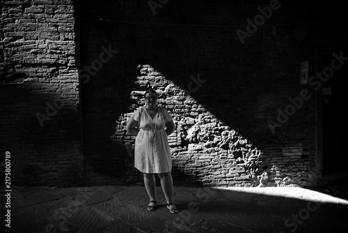 Bild auf Leinwand Woman Standing in Triangular Beam of Sunlight Against Brick Wall in High Contras