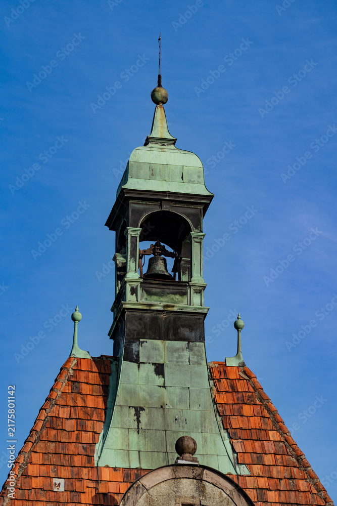 Fototapeta premium Vintage bell tower with green copper patina roof on old building. Ancient church or town hall bell tower featuring weathered copper elements and orange clay roof tiles under blue sky