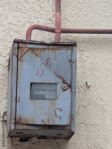 An old electricity meter hanging on a wall, supported by rusted pipes. Faded dial, peeling paint, and years of neglect.

