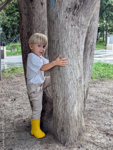 Bild auf Leinwand Toddler in yellow rain boots peeking from behind a tree in the park