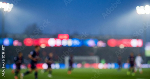 A blurry action shot from a soccer match at dusk, showcasing players on the field under stadium lights with an audience in the background.