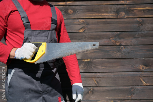 Cuadro en lienzo Red clad worker holding handsaw in protective gloves and dark work overalls near