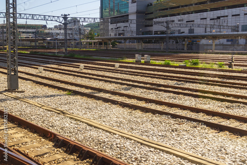 Foto Multiple railway tracks run past station platforms and modern urban buildings beneath a dense network of overhead electrical wires