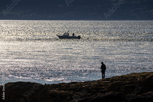 Canvastavla Silhouetted angler fishing from rocky shore with boat on shimmering water at Pen