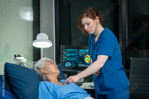 Nurse in blue scrubs checks an elderly female patient with a stethoscope in a modern hospital room, with medical monitors and clinical equipment in the background.