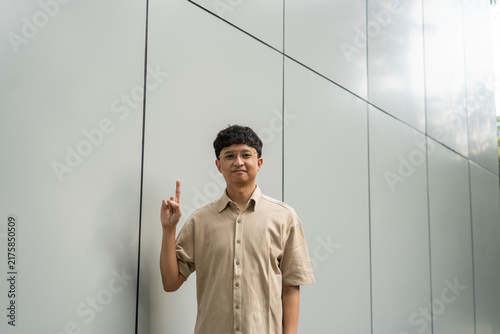 Konstfotografi A young man stands in front of a modern building, raising his finger in a gesture of