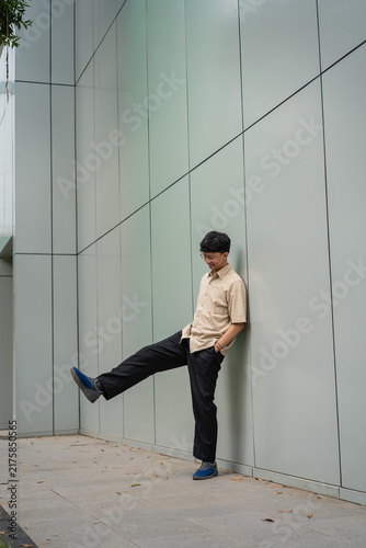 Fotografie A young man in a beige shirt casually leans against a modern gray wall outdoors