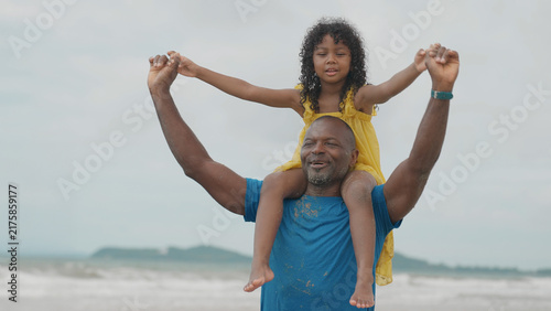 Foto A Black father carries his daughter on his shoulders at the beach under an overcast sky