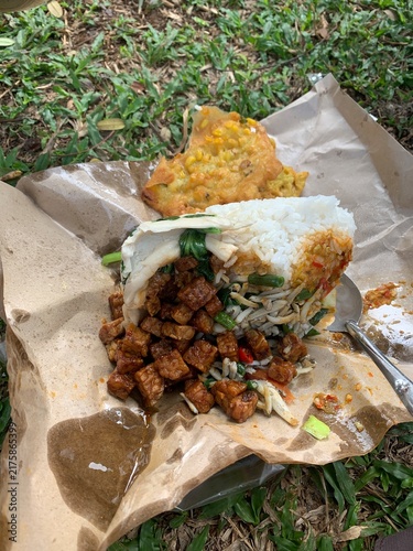 Bild auf Leinwand Authentic Indonesian mixed rice dish served on traditional brown paper wrap on green grass, featuring fried tempeh, stir-fried vegetables, and corn fritter for an outdoor lifestyle meal