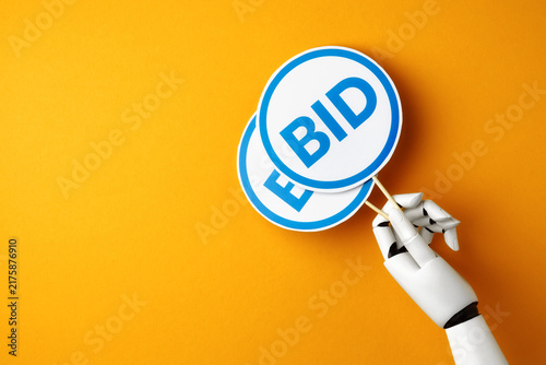 Foto A white robotic hand holding a two blue bidding paddle with BID signs on a bright yellow background