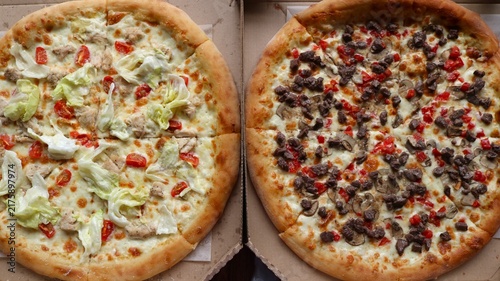 Two round pizzas in open boxes. On the left is a light pizza with chicken, cherry tomatoes, and lettuce, reminiscent of a Caesar salad. On the right is a hearty pizza with beef and mushrooms.