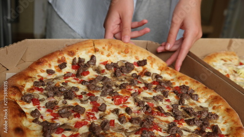 A close-up shows an appetizing pizza in an open box. The thick, golden-crusted dough is generously topped with juicy pieces of meat, sliced ​​mushrooms, tomatoes, and melted cheese.