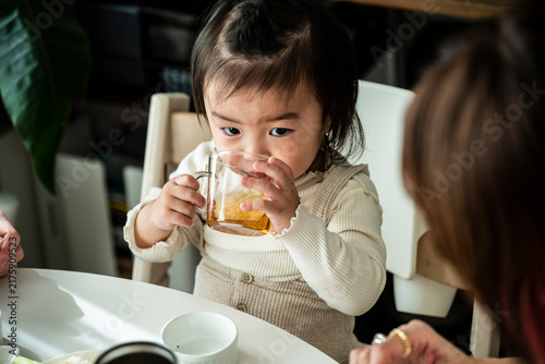 Cute Asian Toddler Girl Drinking Tea from Glass Cup at Dining Table with Family
