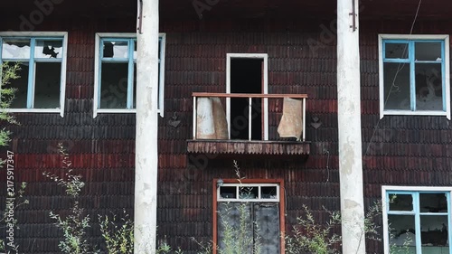 Static view of a weathered wooden building featuring broken windows, a rusted balcony, and overgrown vegetation, maintaining a consistent unmoving perspective of the deteriorating structure.