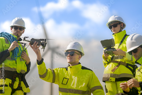Bearded technician at the left of frame steadies a quadcopter while the fair haired pilot beside him reaches up and a blonde colleague holds the controller at the right