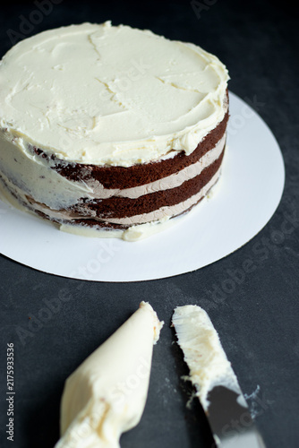 Chocolate layer cake with white frosting on a round cake board, spatula with frosting placed beside it
