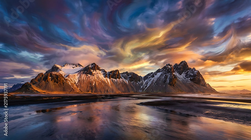 Dramatic Mountain Sunset: Jagged, snow-capped mountain peaks are silhouetted against a sky ablaze with fiery orange, purple, and yellow clouds, reflected in the calm, dark water below.