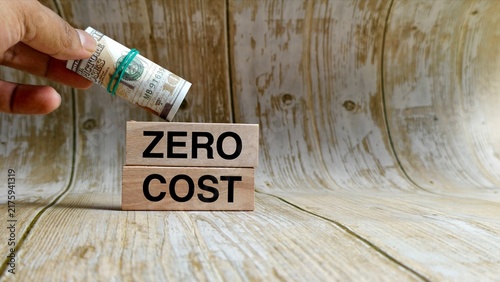 Hand holding a rolled US dollar bill over wooden blocks reading ZERO COST on a wooden surface.