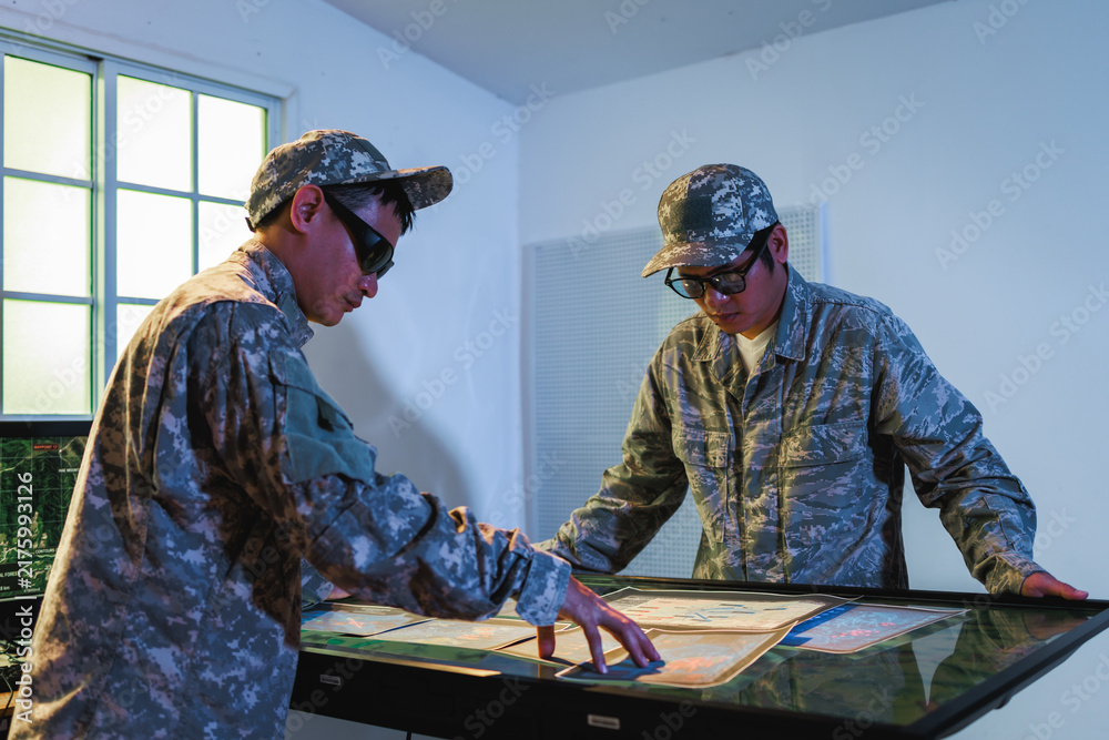 Naklejka premium Two uniformed military personnel study maps on a lighted planning table inside a command room, preparing for a mission.