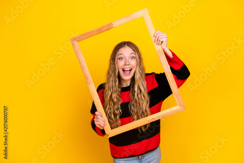 Obraz na plátně Cheerful young woman holding a wooden frame against a bright yellow background,