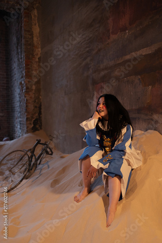 Fotografie Circus performer in blue pierrot costume sitting barefoot on sand beside an old