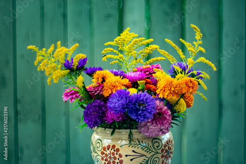 A bouquet of bright autumn flowers in a hand-painted ceramic vase against a green wooden wall.