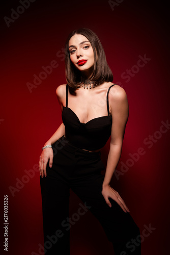 Glamorous young woman in a black evening outfit posing against a red background with bold red lipstick and statement jewelry